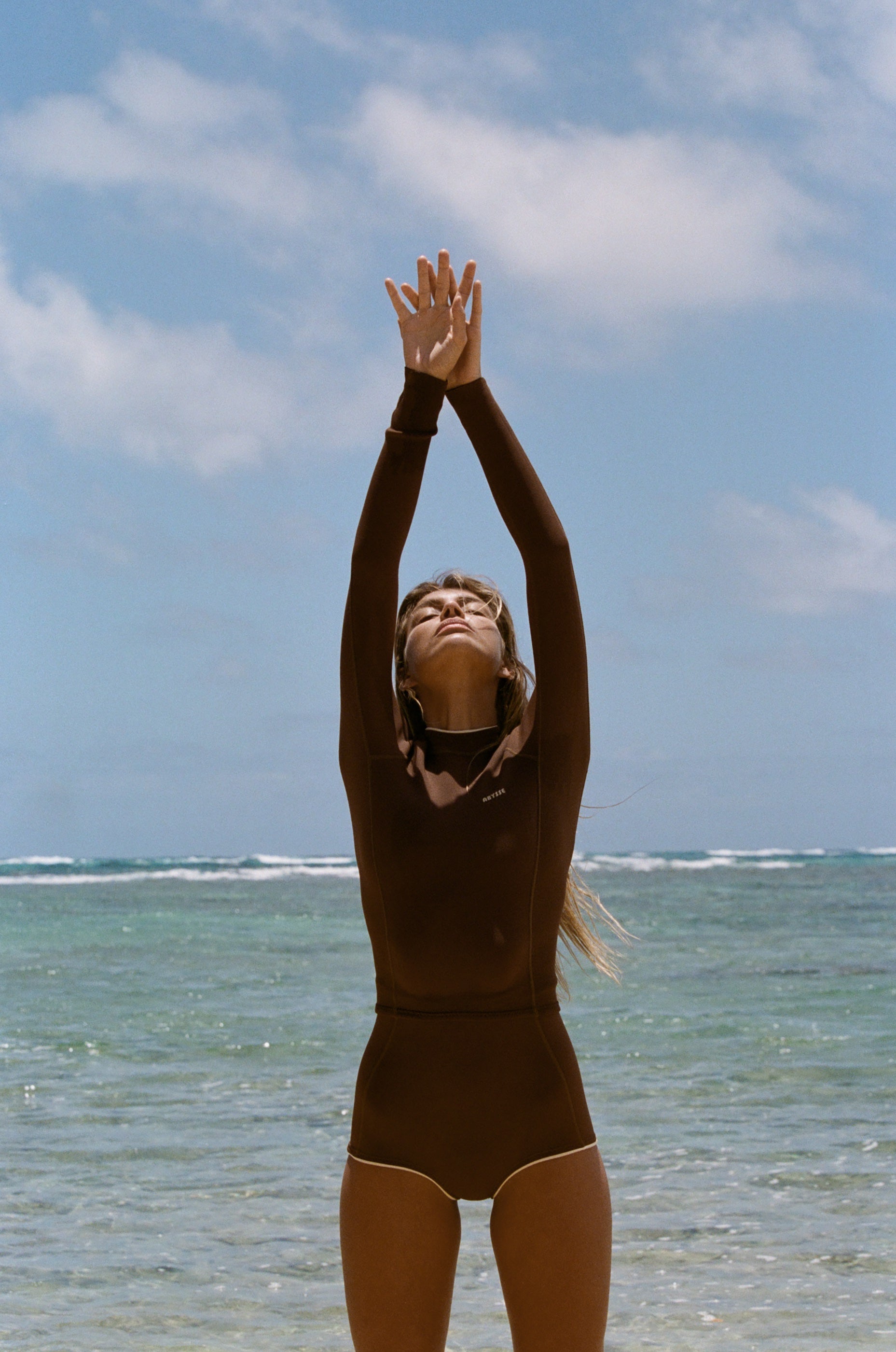 A woman in a dark brown full-body swimsuit or wetsuit stands on a beach with her arms raised above her head, looking upward, with the ocean and blue sky in the background.