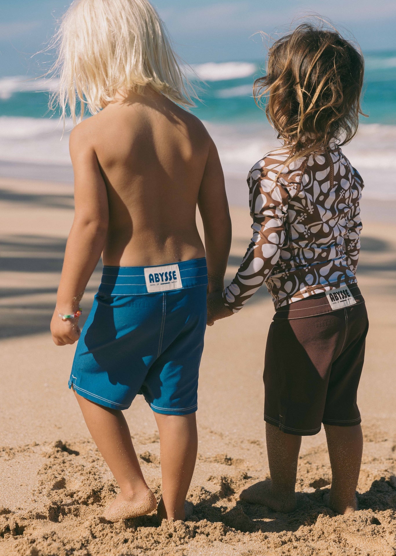 Two children holding hands on a sandy beach with ocean waves in the background.