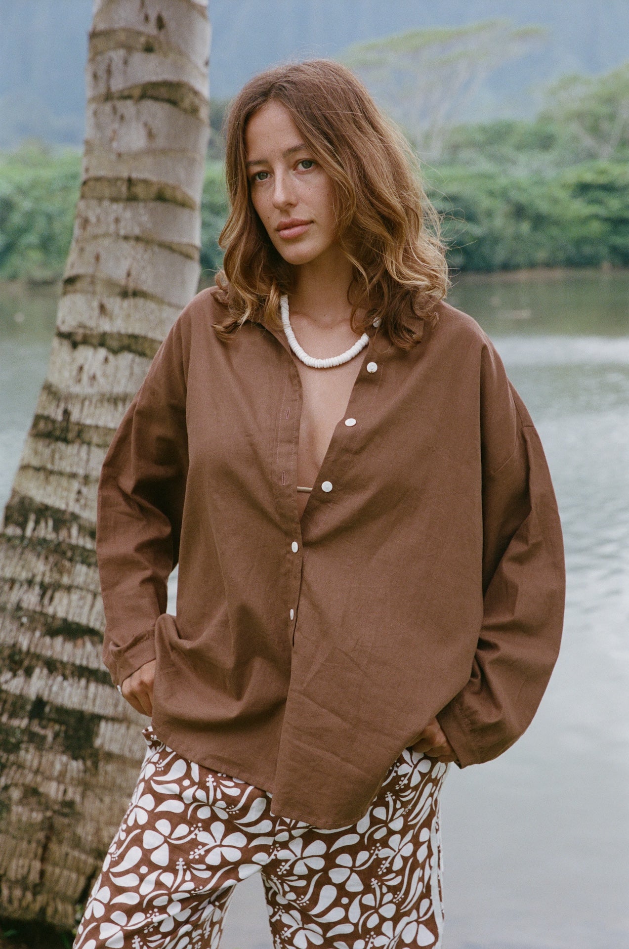 Woman wearing a brown jacket and patterned pants standing by a body of water with trees in the background