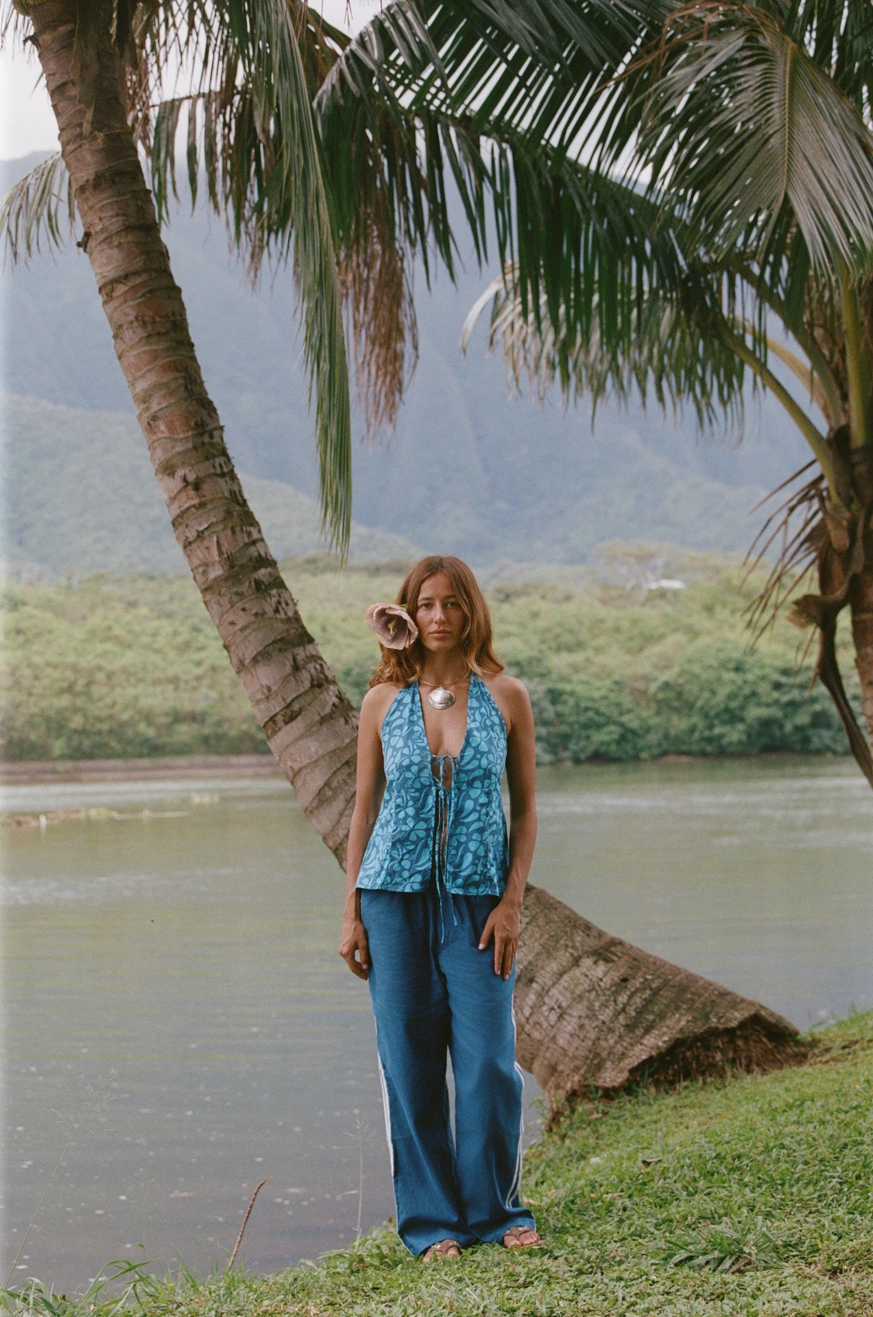 Woman in a blue top and pants standing by a palm tree by a body of water with mountains in the background.