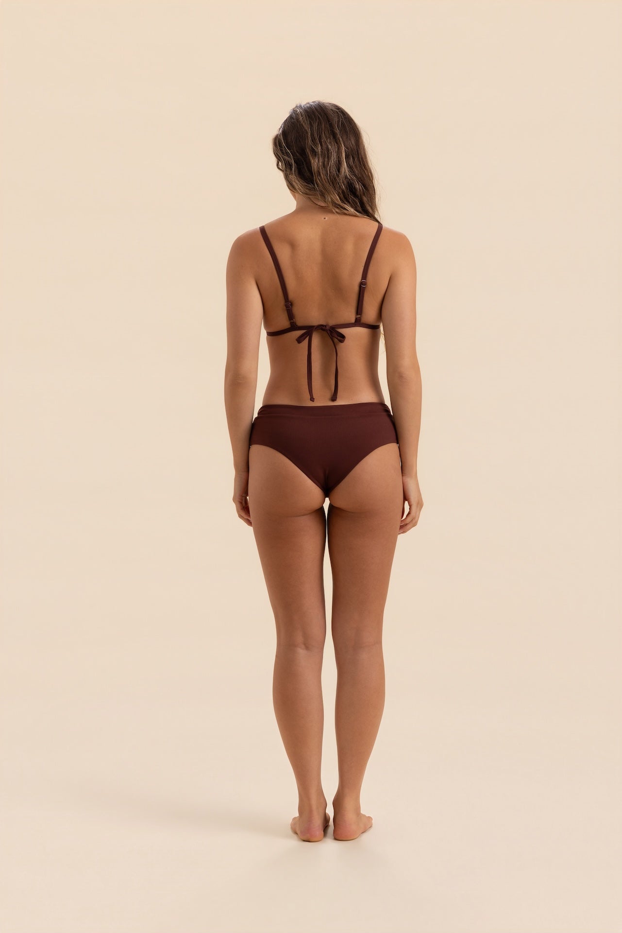 A woman viewed from behind wearing a dark brown string bikini against a solid beige studio background.