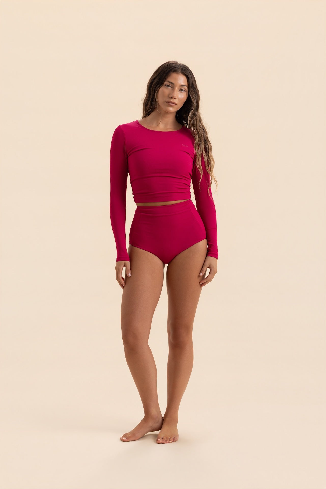 A woman modeling a bright magenta two-piece swimsuit consisting of a long-sleeve crop top and high-waisted bottoms, standing against a solid beige background.
