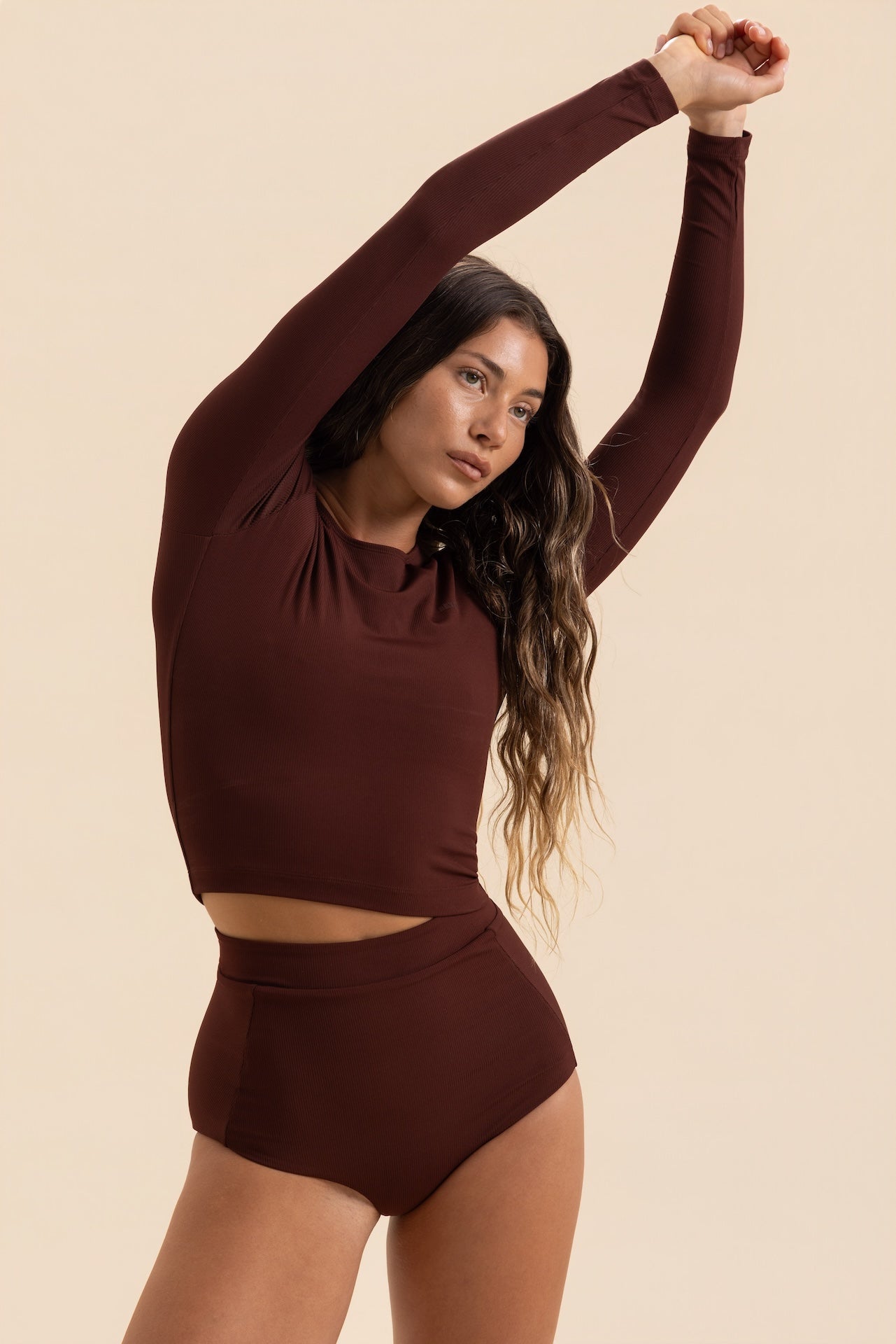 A woman modeling a matching two-piece activewear set in a deep burgundy color, featuring a long-sleeve crop top and high-waisted shorts, against a solid beige background.