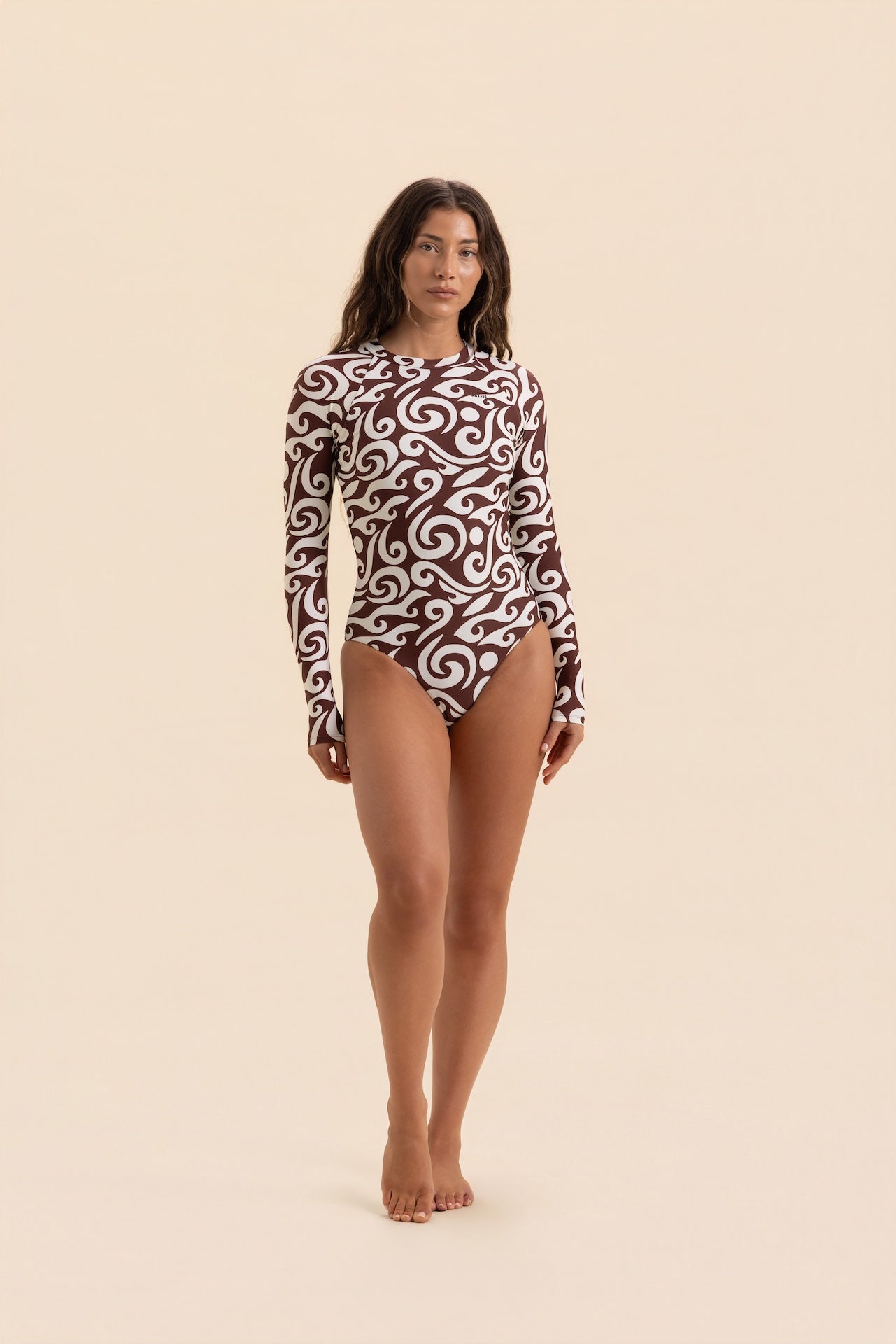 A woman modeling a long-sleeve one-piece swimsuit with a brown and white swirl pattern, standing against a solid beige background.