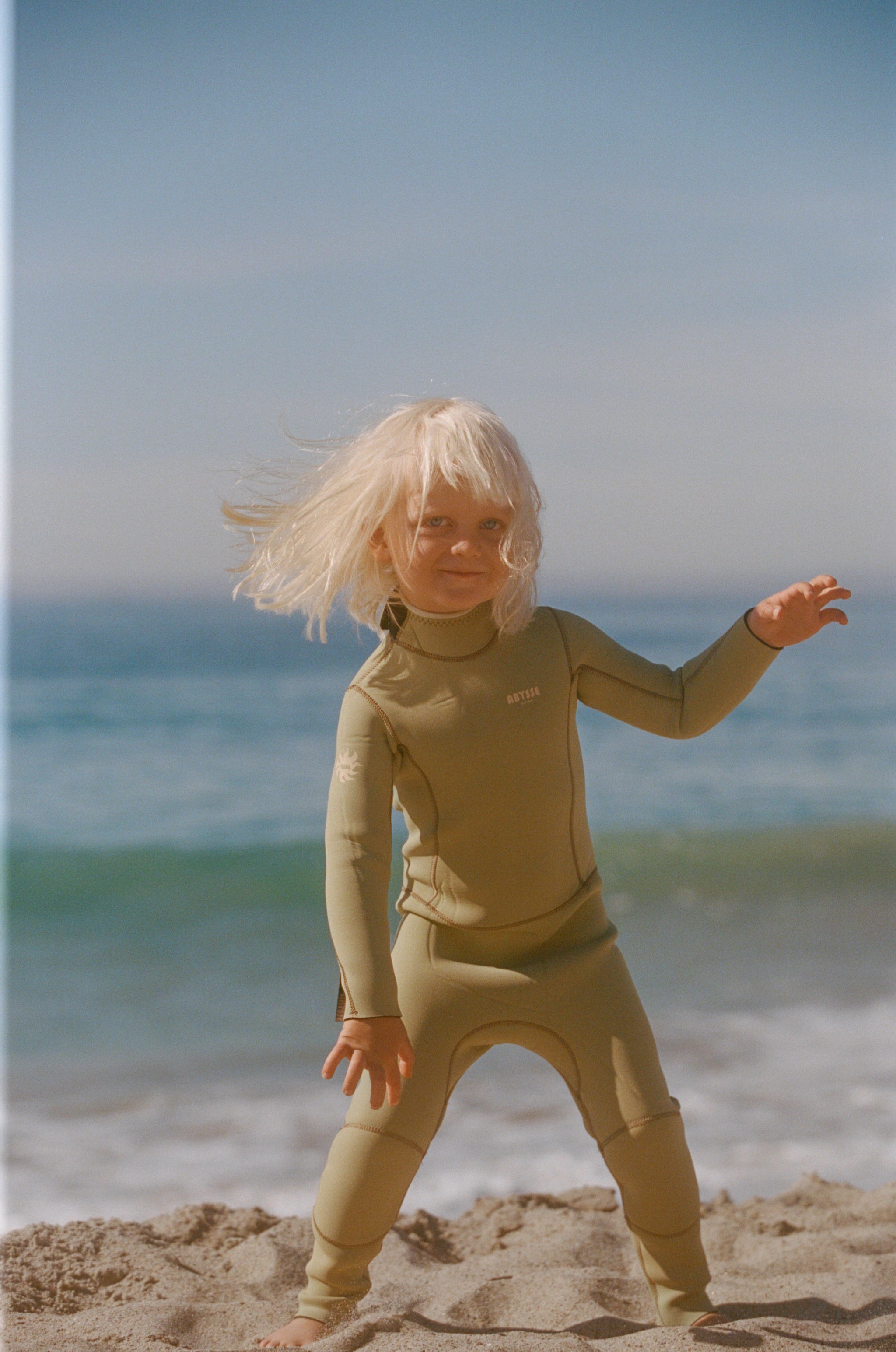 Child in a wetsuit standing on a beach with ocean in the background
