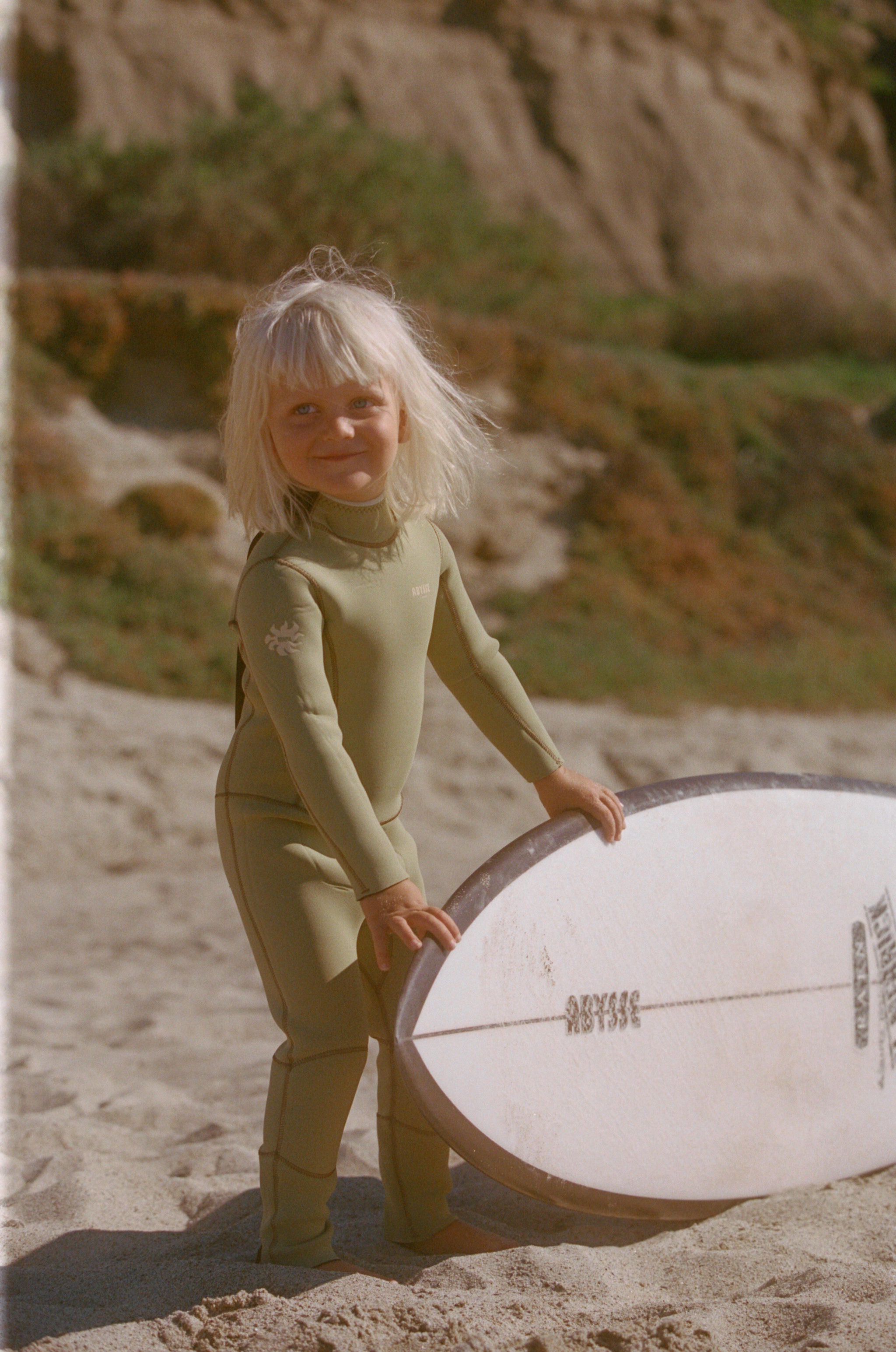 Child in a green wetsuit holding a white surfboard on a sandy beach.