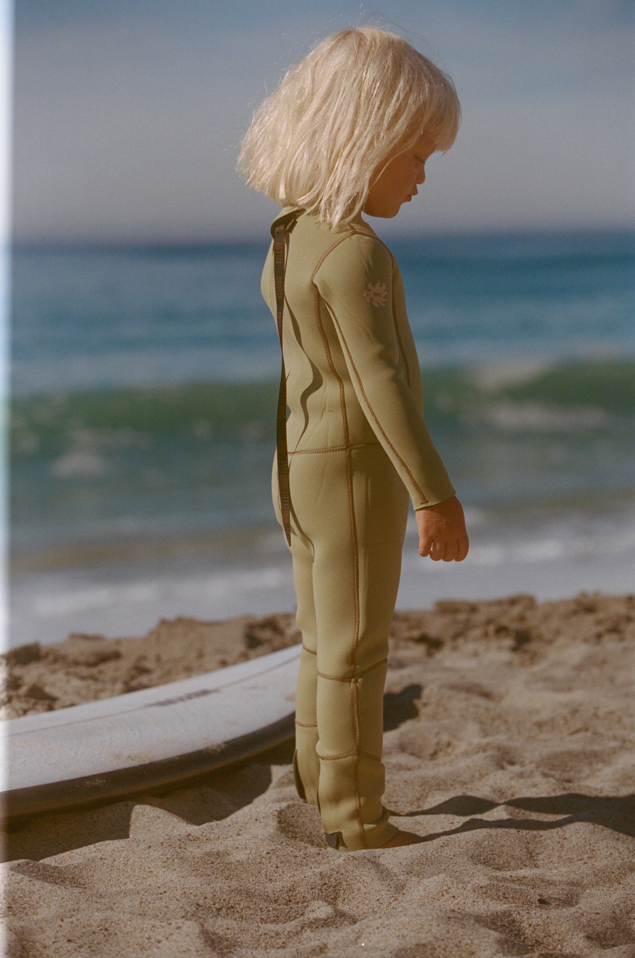 Child in a green wetsuit standing on a sandy beach with ocean waves in the background
