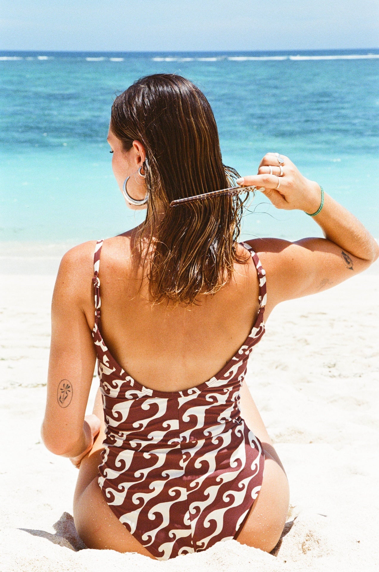 Woman in a patterned swimsuit sitting on a beach with clear blue water.