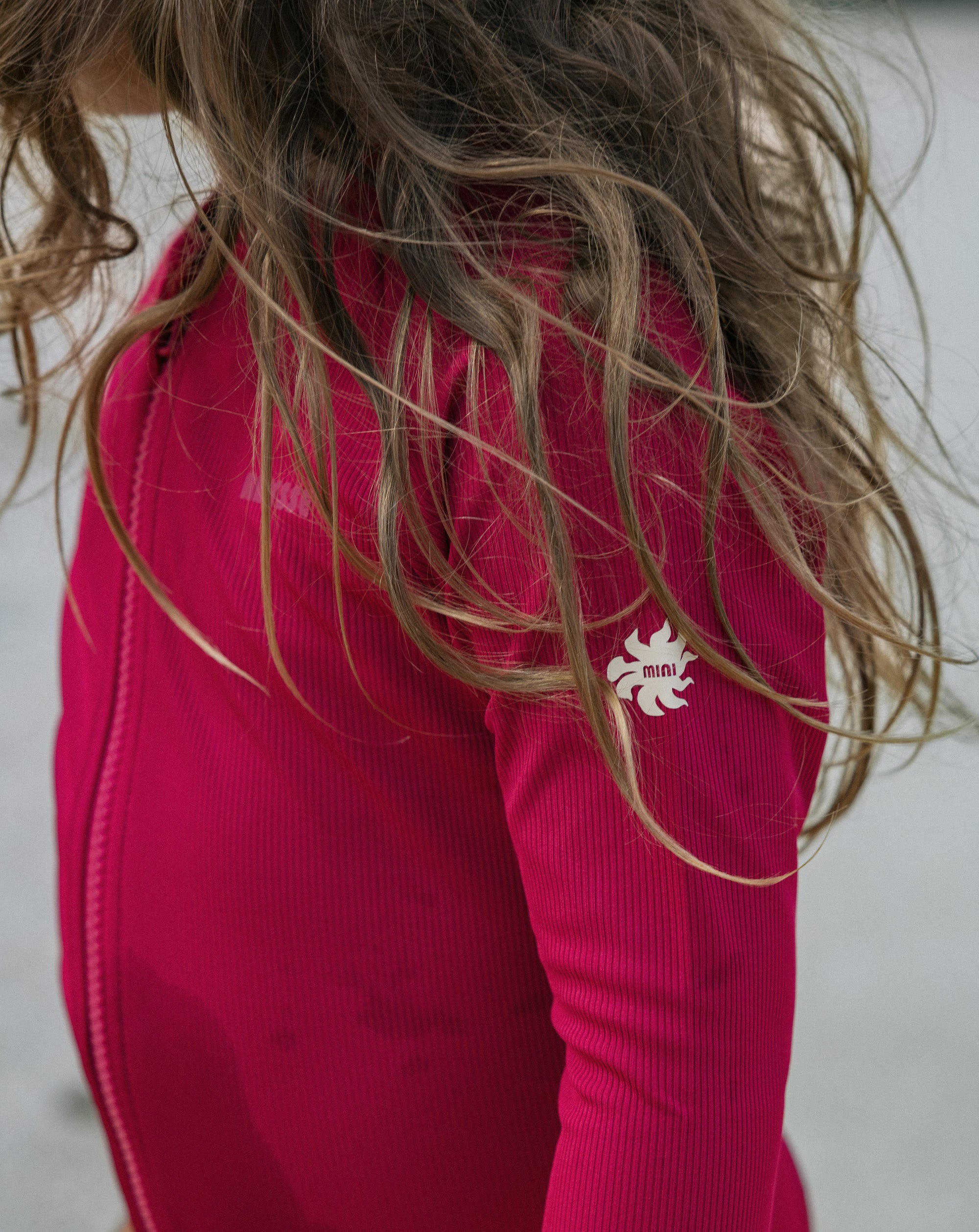 A close-up of a child wearing a bright pink ribbed long-sleeve top, with long light brown hair blowing around. A small white stylized logo is visible on the upper sleeve.