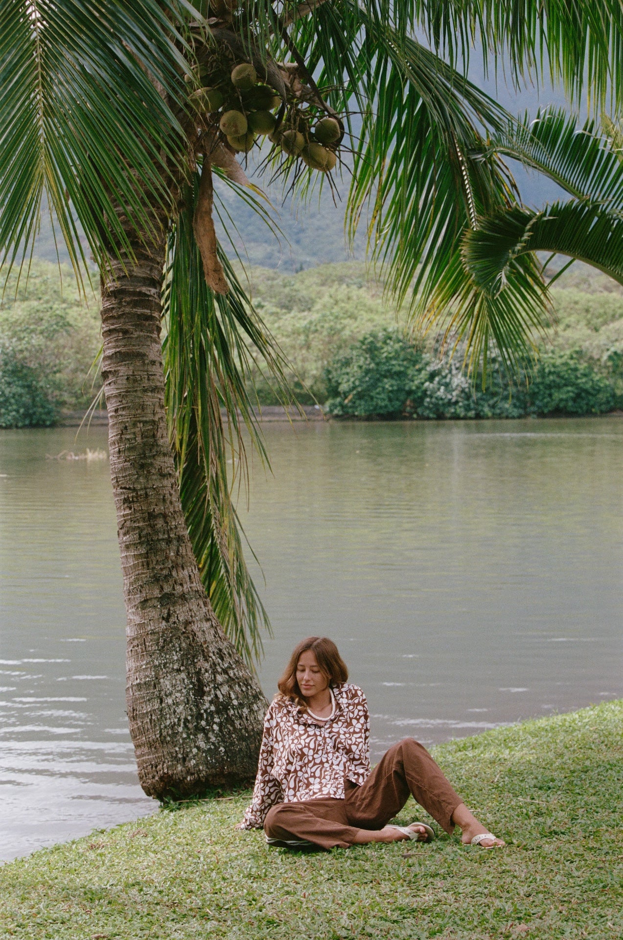 Woman sitting under a palm tree by a body of water with mountains in the background