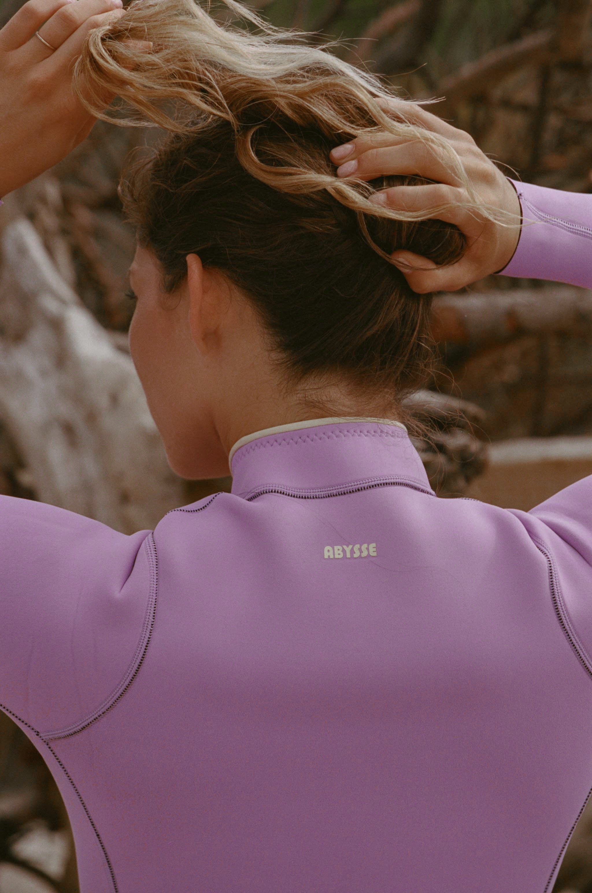 A woman wearing a lavender-purple wetsuit, seen from behind as she adjusts her hair. The brand name 'ABYSE' is printed in small gold letters on the back of the wetsuit collar.