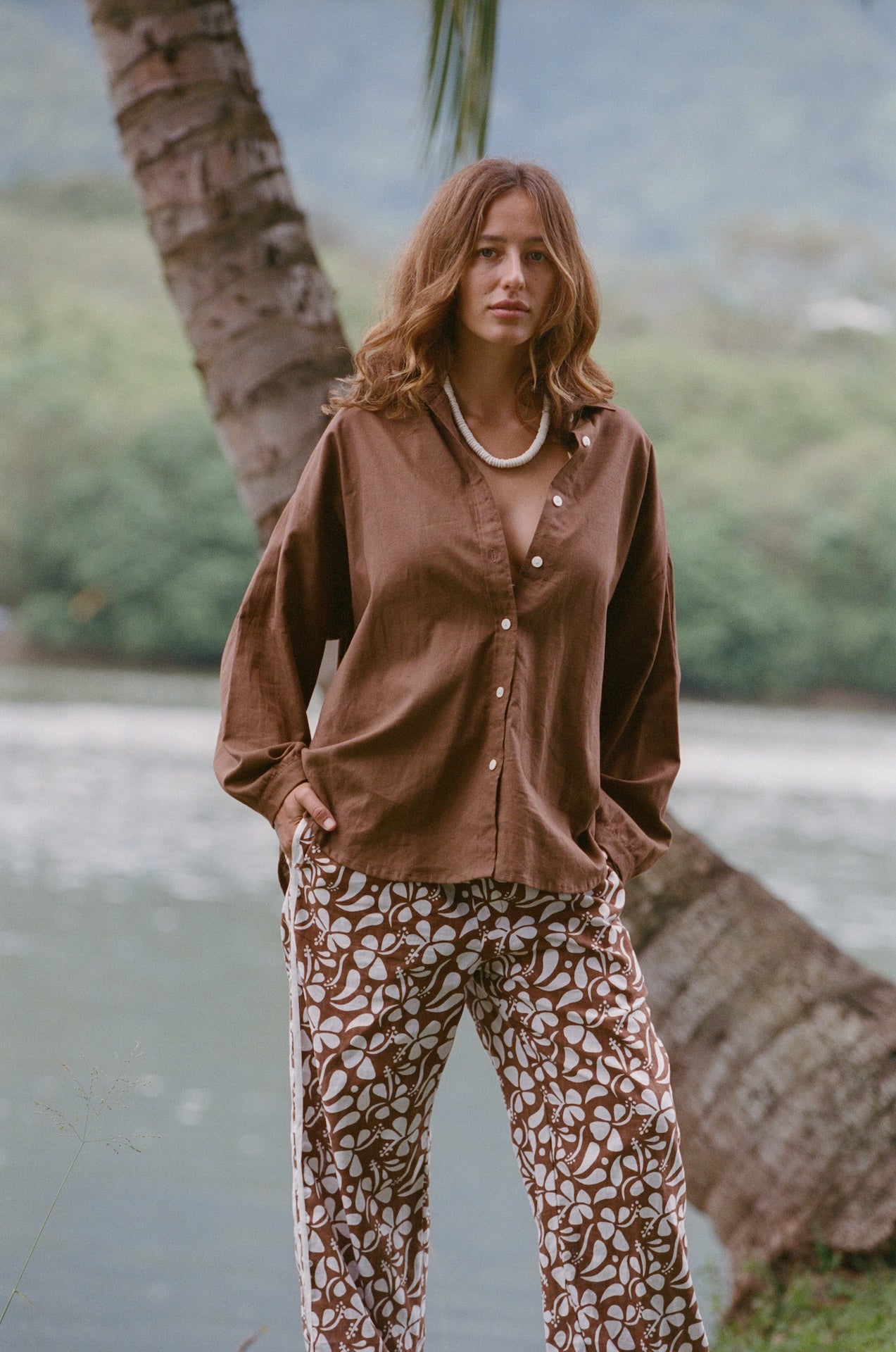 Woman wearing a brown shirt and patterned pants standing next to a palm tree