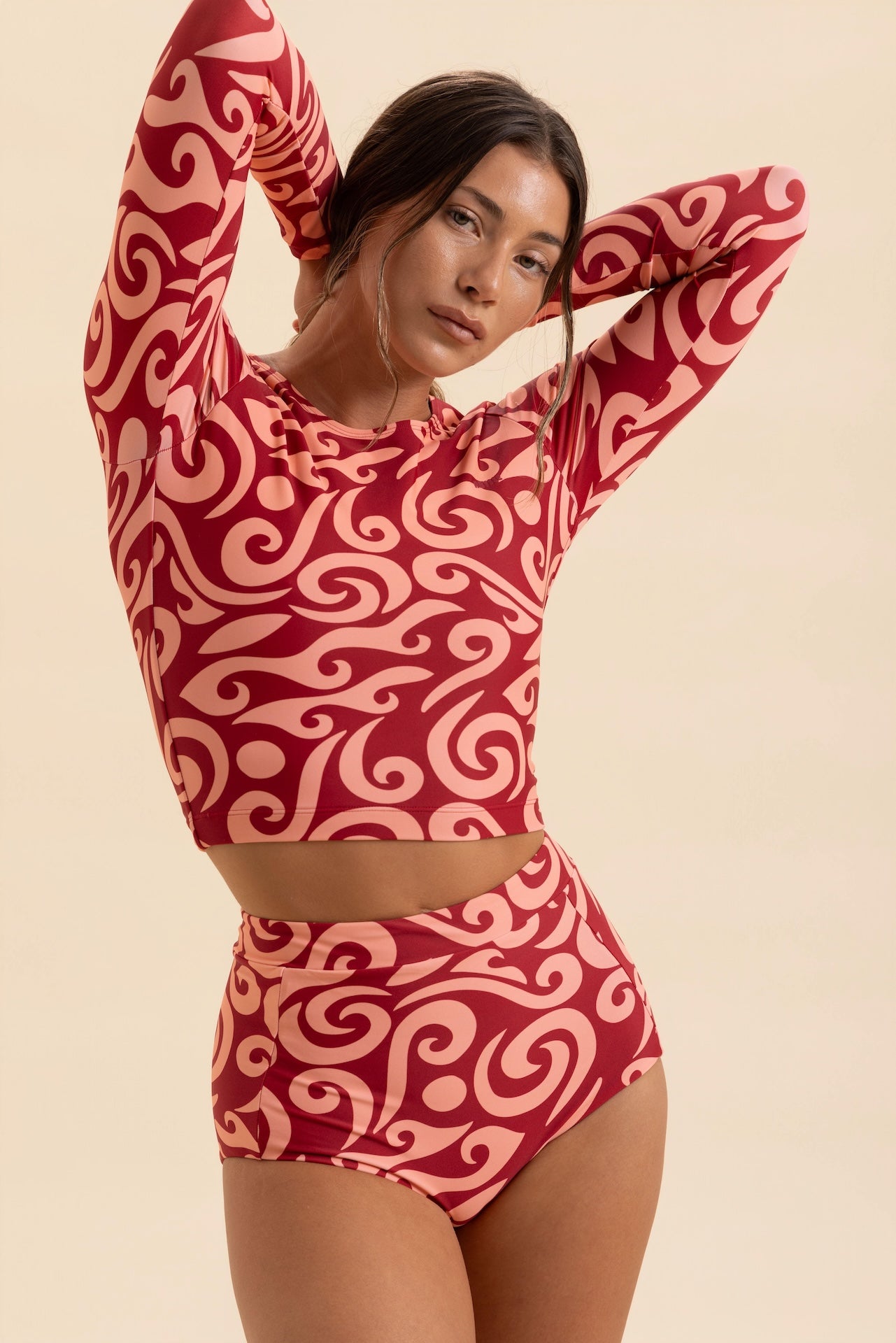 A woman modeling a matching two-piece swimsuit with a red and pink abstract swirl pattern, featuring a long-sleeve crop top and high-waisted bottoms, against a solid beige background.