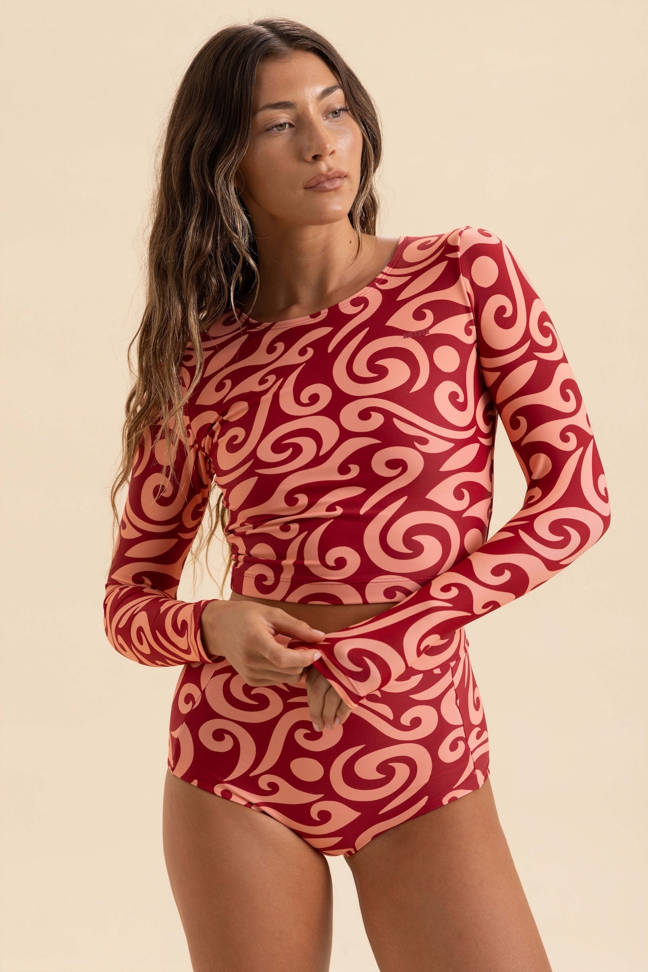 A woman modeling a long-sleeve one-piece swimsuit with a red and light pink abstract swirl pattern, set against a solid beige background.