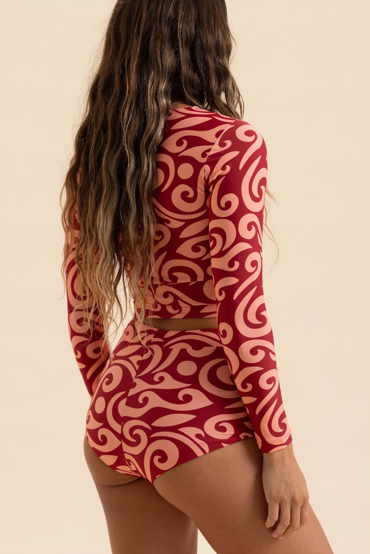 A woman shown from the back wearing a long-sleeve red and pink patterned crop top and matching high-waisted shorts against a solid beige background.