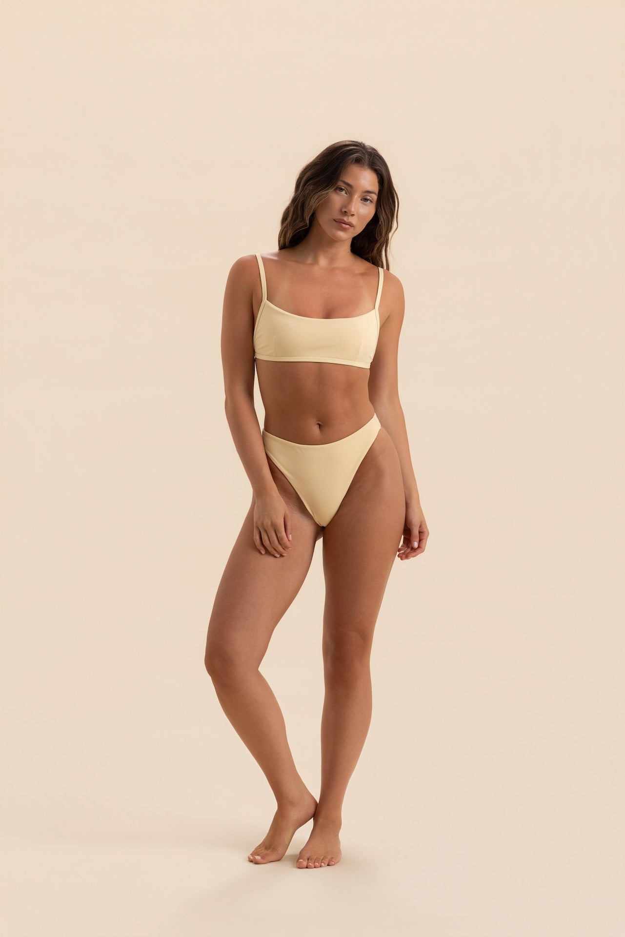 A woman modeling a two-piece bikini swimsuit in a solid pale cream color, standing against a matching solid cream background.