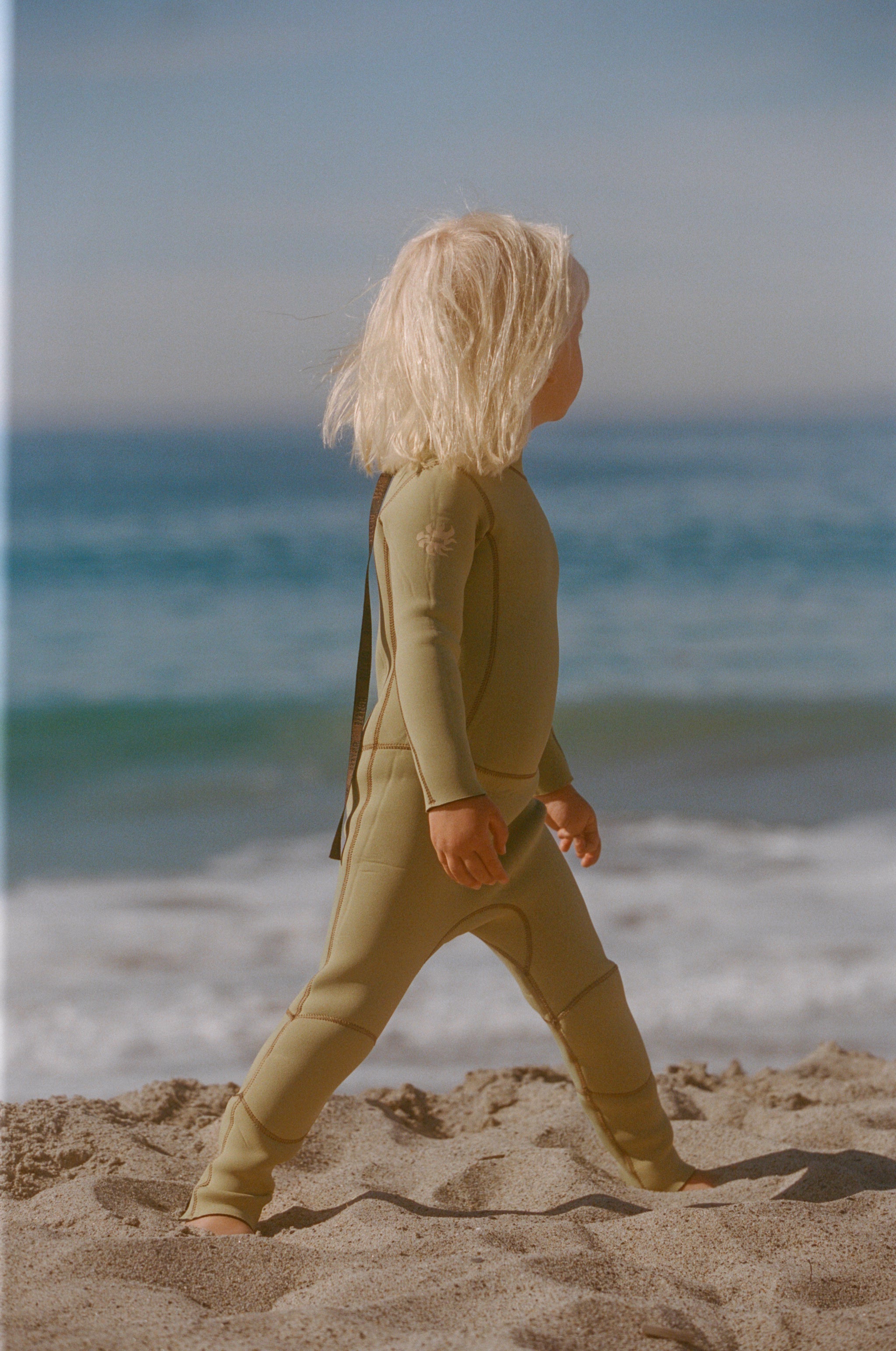 Child in a green suit walking on a sandy beach with ocean waves in the background
