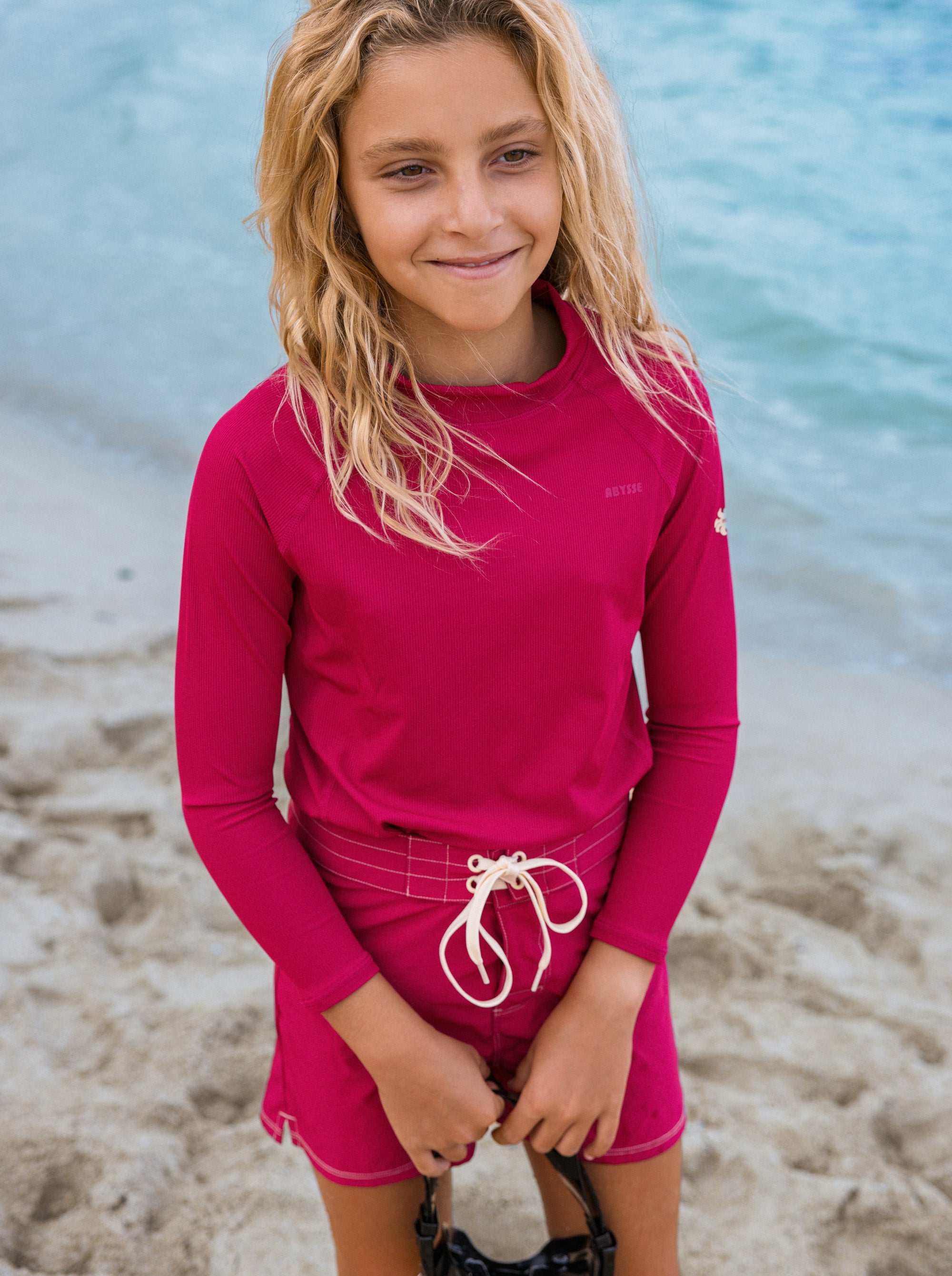 A young girl with blonde hair stands on a sandy beach wearing a bright pink long-sleeve rash guard and matching pink swim shorts with a white drawstring.