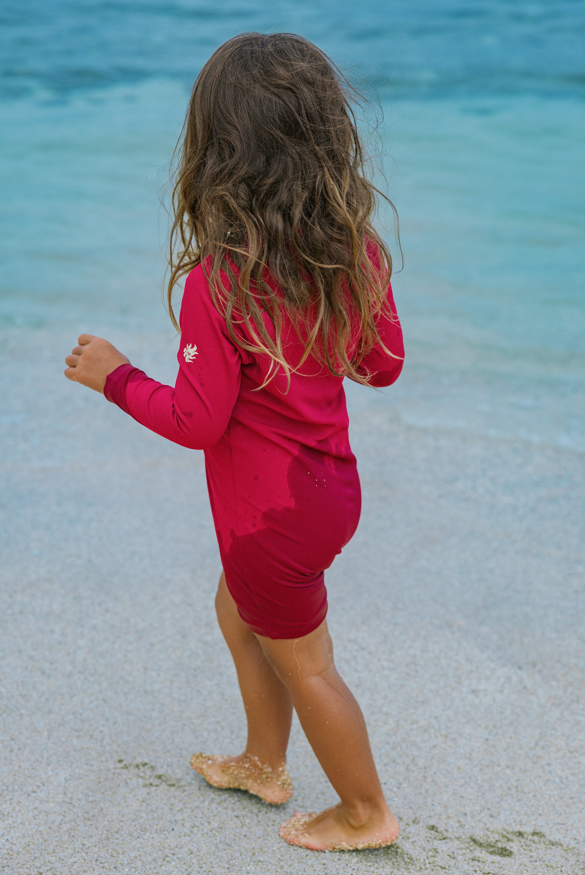 A young girl with long wavy brown hair stands on a sandy beach facing the ocean, wearing a bright magenta-pink long-sleeve rash guard and matching swim shorts.