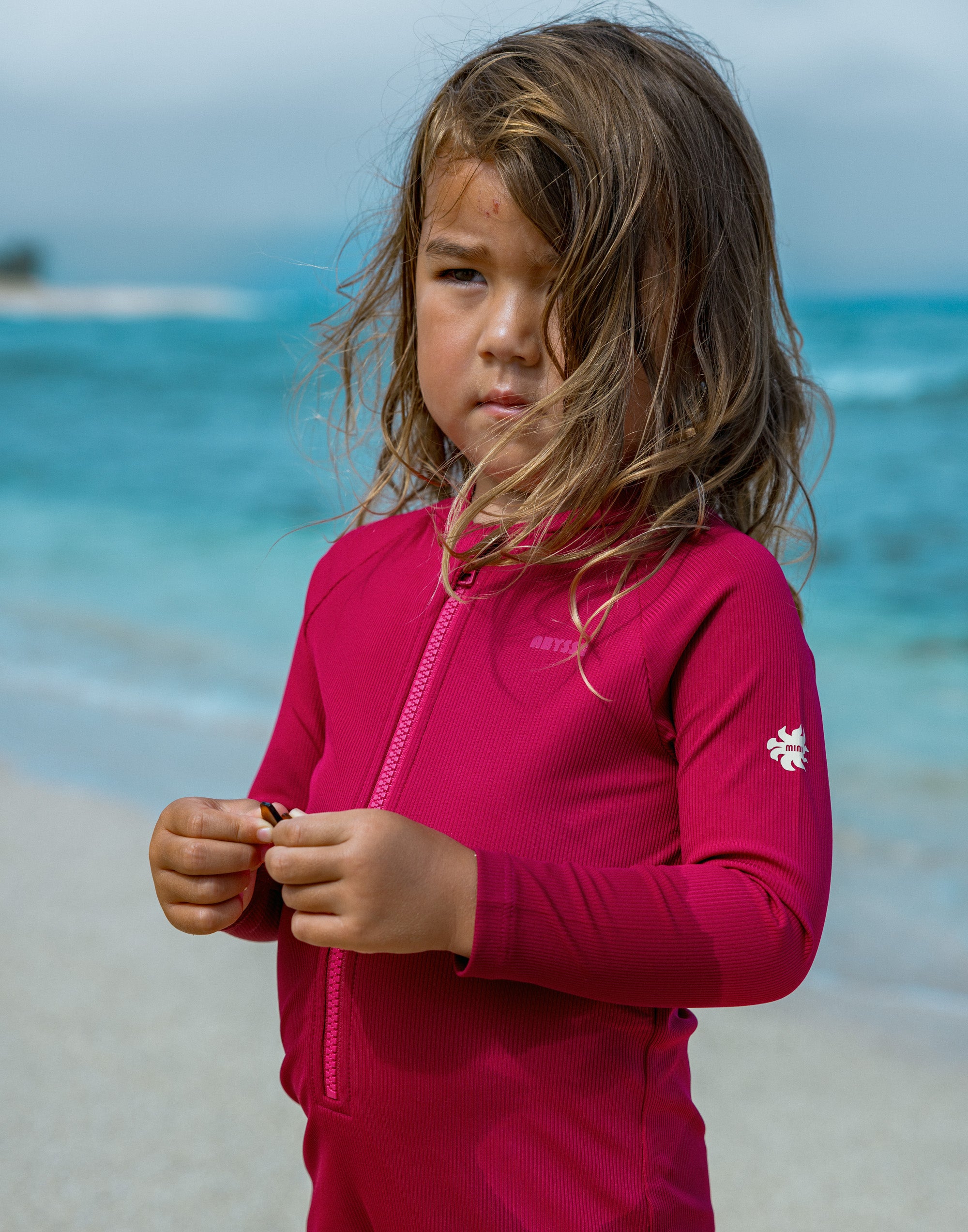A young girl with long brown hair stands on a beach wearing a bright pink long-sleeve zip-up rash guard. The brand name 'Antifreeze' is visible on the chest, and a small logo is on the sleeve.