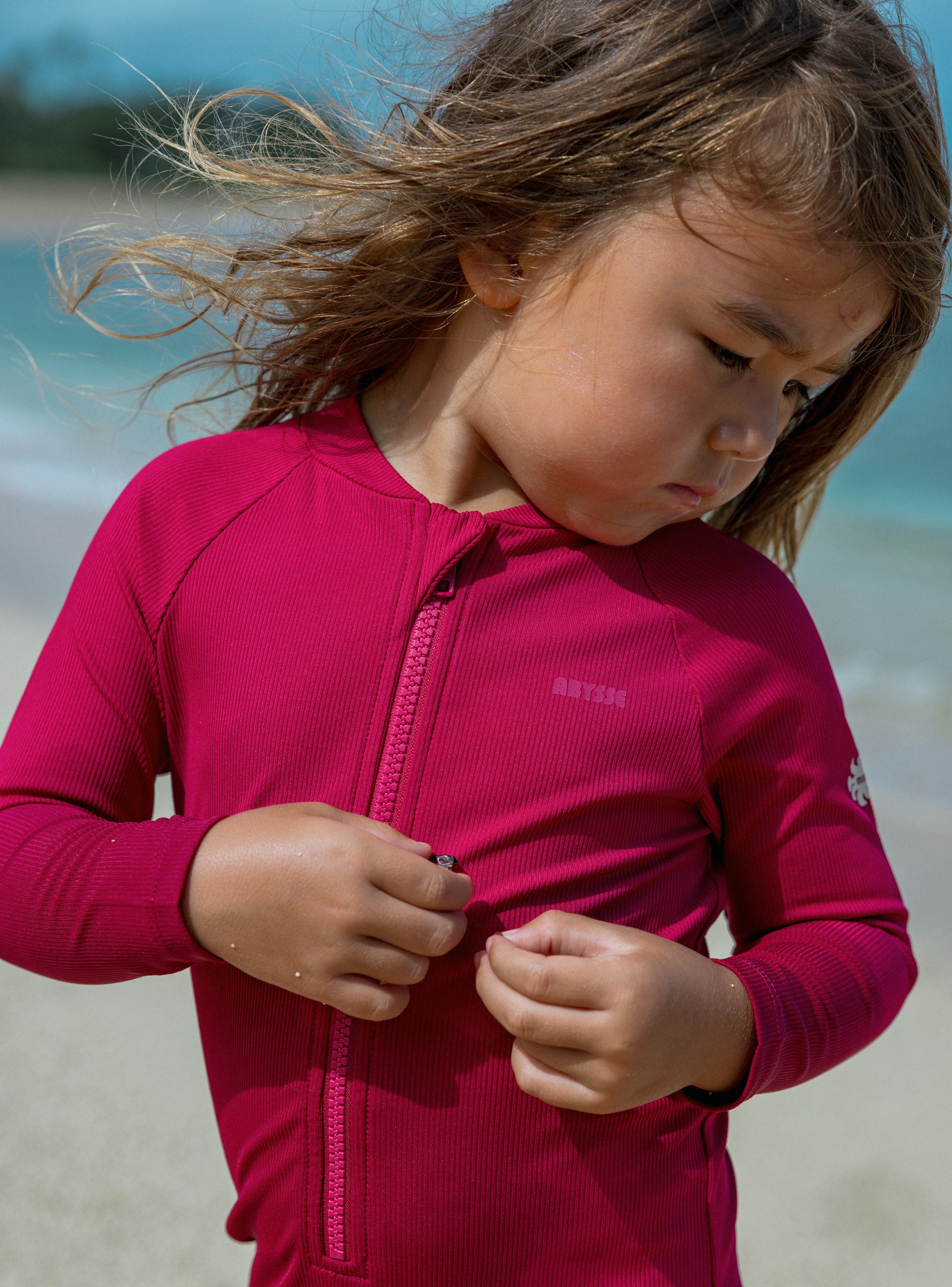 A young girl with windblown brown hair looks down while zipping up a long-sleeve pinkish-red rash guard on a beach. The brand name 'ATKIM' is visible on the chest.