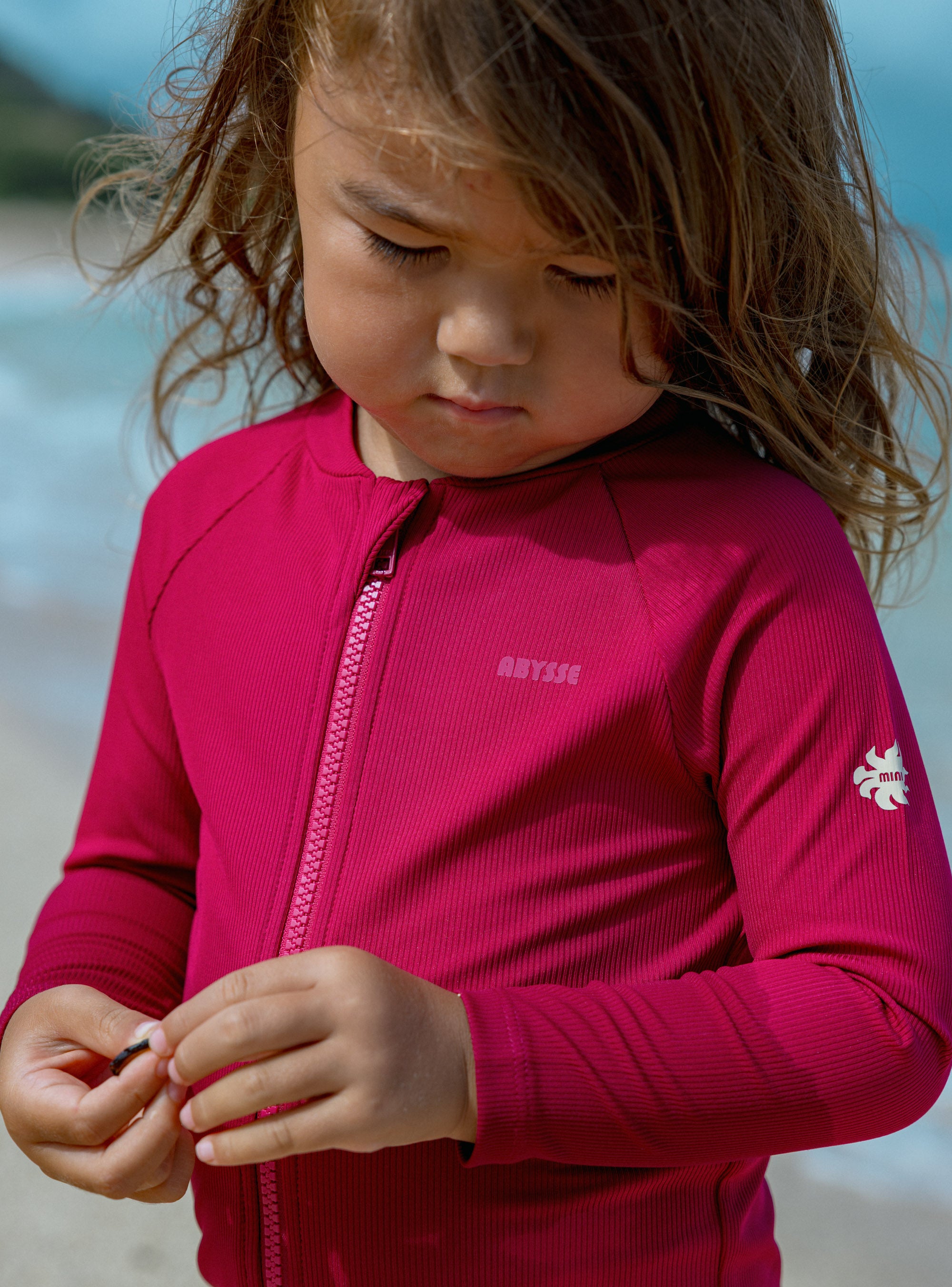 A young child wearing a bright pink Oyster Reef long-sleeve zip-up rash guard swimsuit, standing on a beach with the ocean in the background.