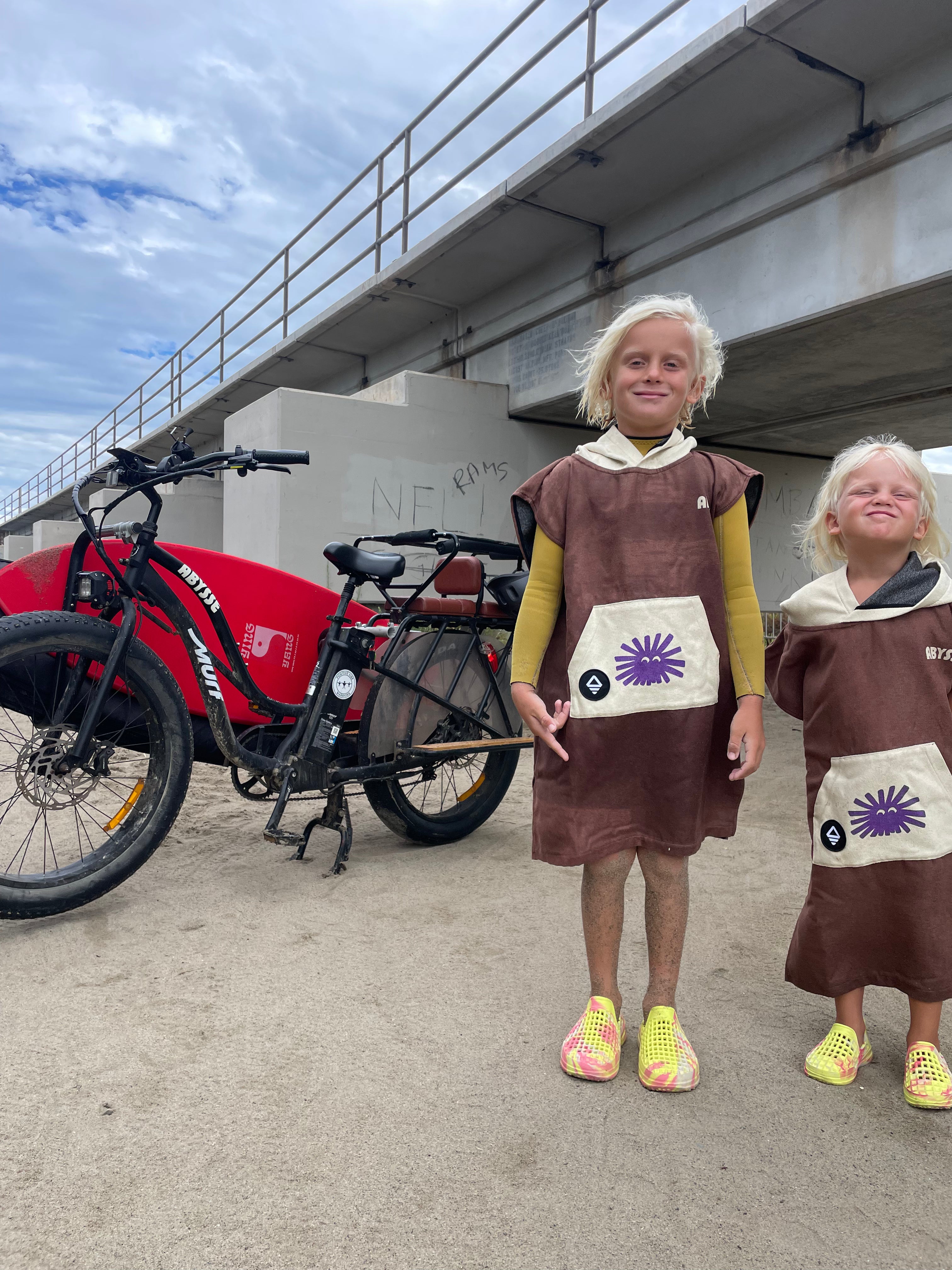 Two children wearing matching brown hooded robes with purple sun patch details stand in front of a red electric cargo bike on an outdoor concrete structure.