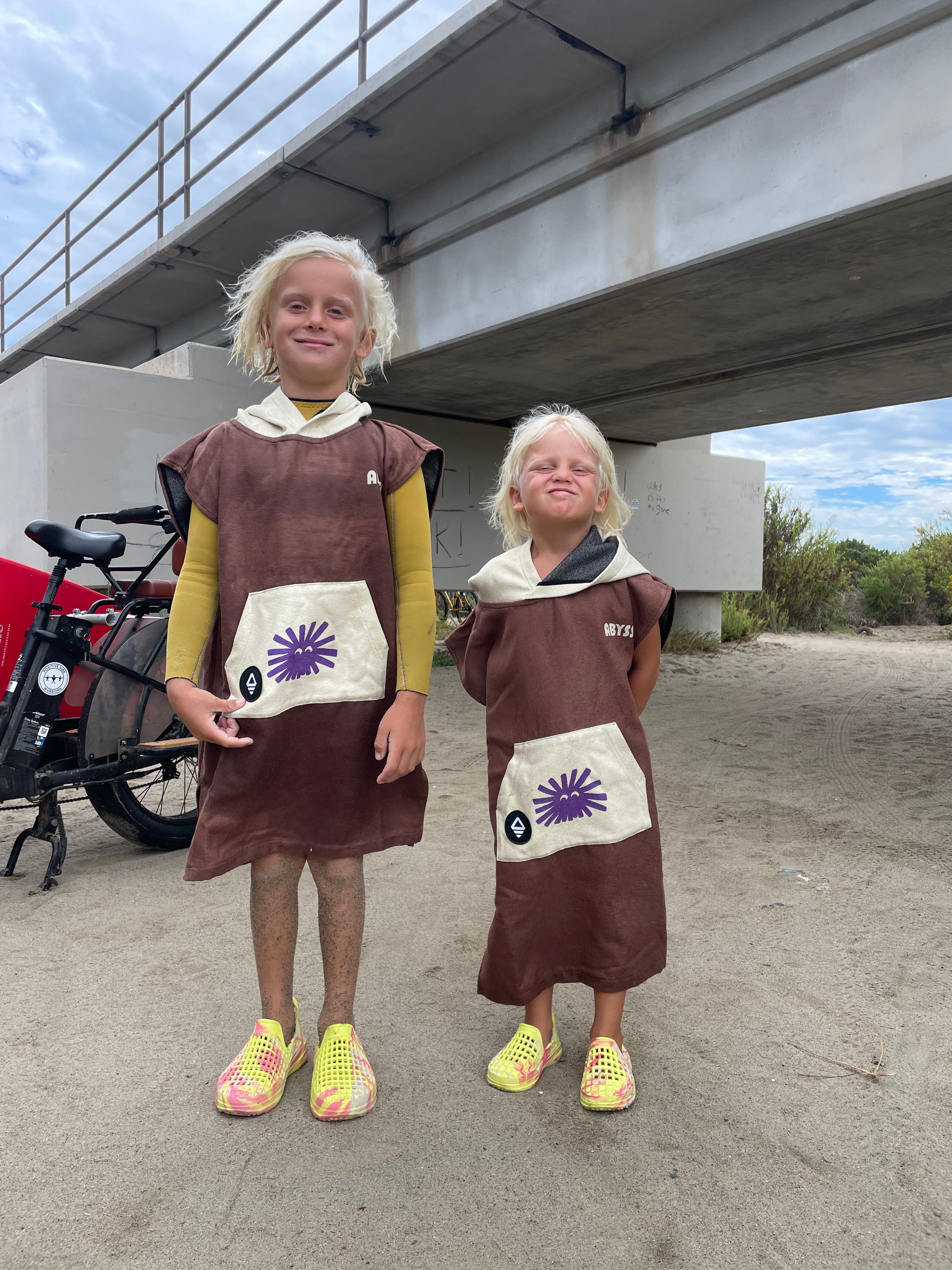 Two children wearing matching brown and cream Star Wars Rebel Alliance hooded robes with purple starburst patches, standing outdoors under a concrete bridge.