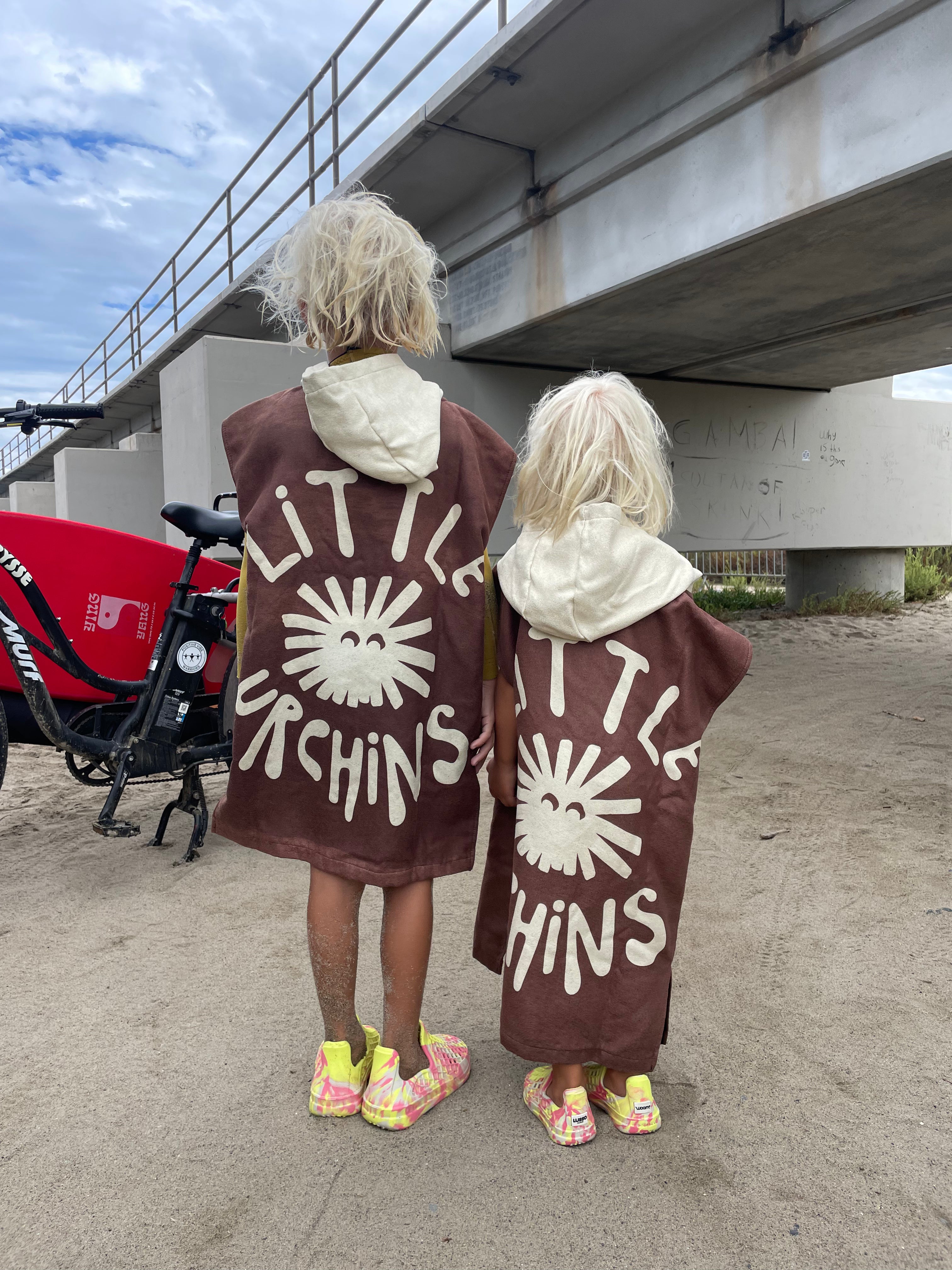 Two children standing from behind under a concrete bridge, wearing matching brown hooded ponchos with a white sun graphic and the text 'LITTLE ORCHINS'.
