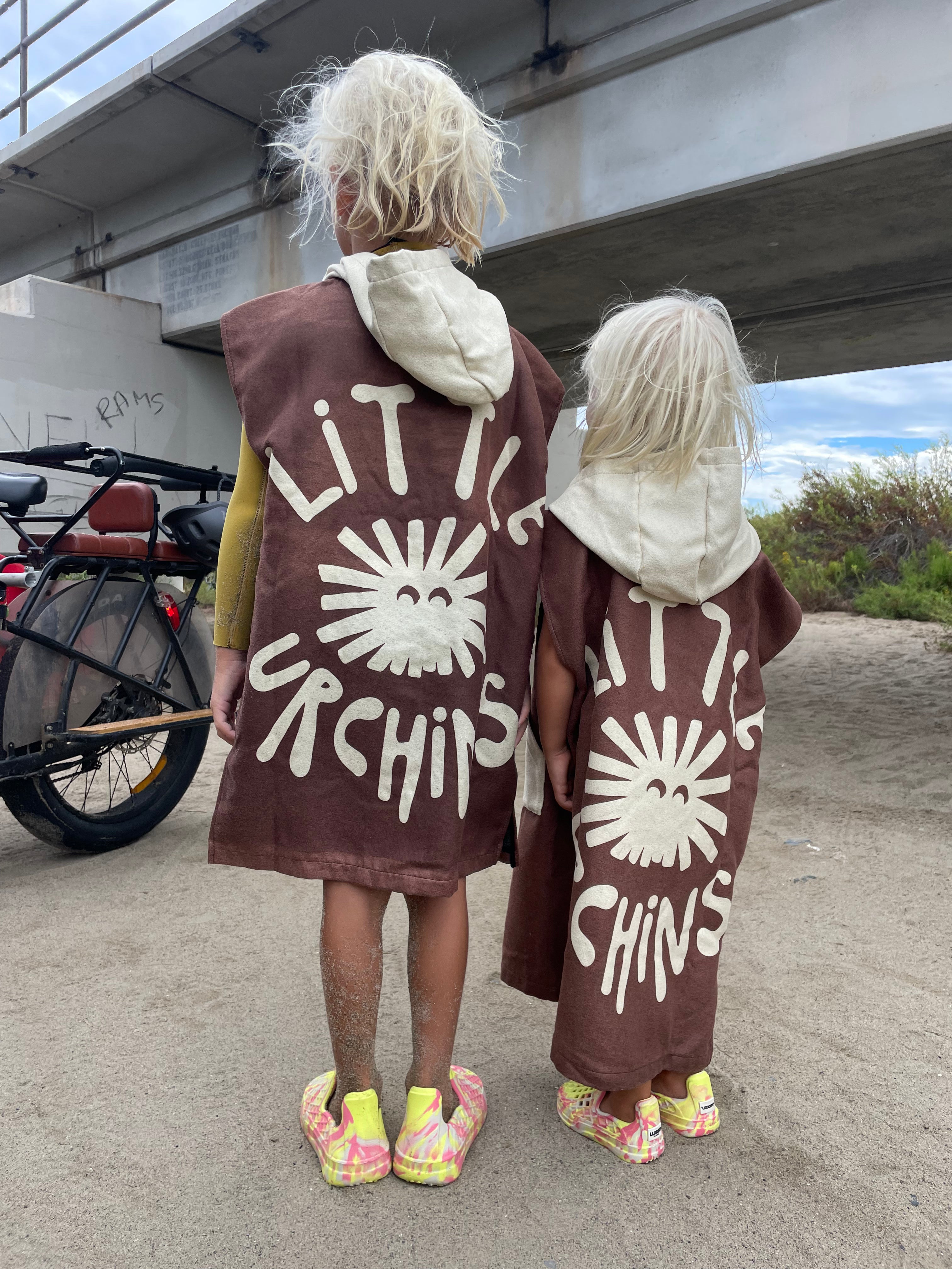 Two children standing outdoors under a bridge, seen from behind, wearing matching brown hooded poncho-style garments with large white 'LITTLE ORCHIDS' text and sunburst graphics.