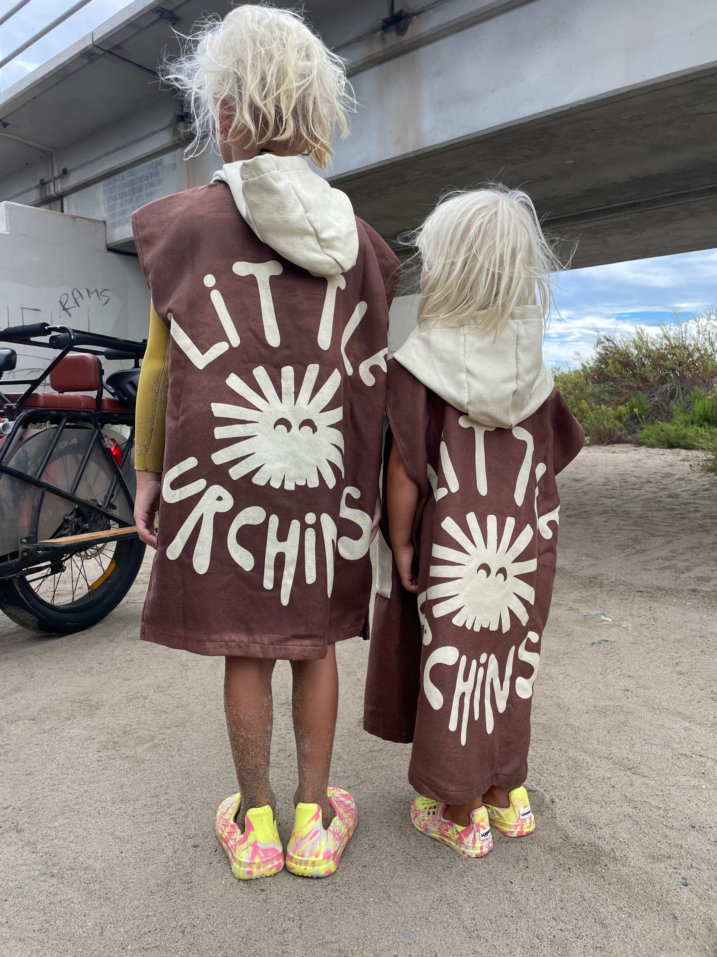 Two children standing outdoors under a concrete bridge, viewed from behind, wearing matching brown hooded cloaks with large white 'LITTLE SPICHIS' text and sun graphics.